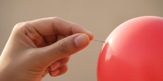 A woman’s hand with natural, unpolished nails holding a small pin against a red balloon, just about to pop it.