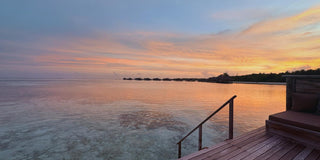 A golden and turquoise sunset over calm Maldivian waters, viewed from a wooden deck with steps leading into the sea at Gili Lankanfushi. The peaceful horizon reflects soft pastel clouds and a feeling of renewal.