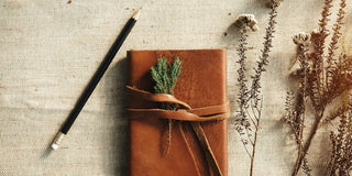 Brown leather journal tied with a leather strap and a sprig of green herbs, beside a pencil and dried flowers on natural linen.