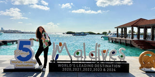 Lisa Eve standing beside the Maldives “World’s Leading Destination” sign at Malé harbor, turquoise water and boats behind her under a bright blue sky.